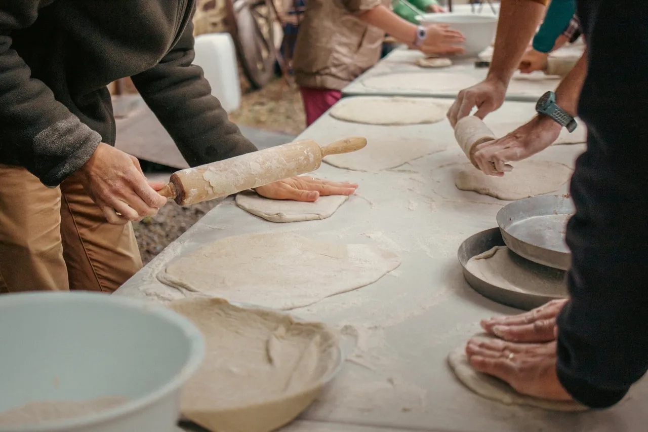 Des mains étalent des pâtes à pizza sur une longue table farinée