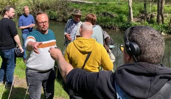 Un groupe de personnes discute au bord d’un cours d’eau ; au premier plan, un homme porte un casque audio