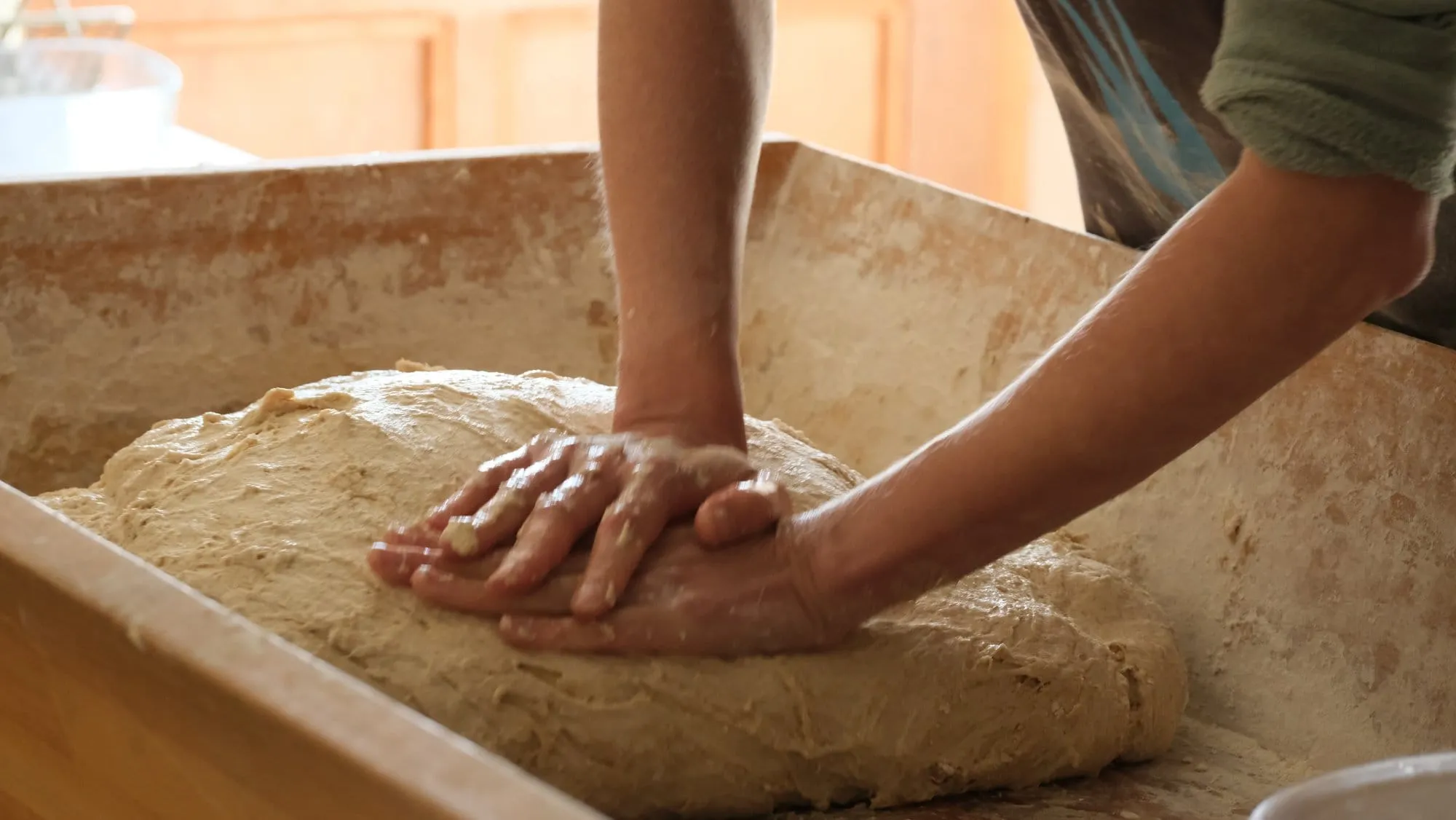 Des mains pétrissent une grosse boule de pâte dans un pétrin en bois